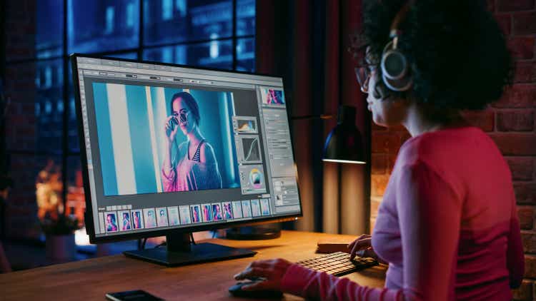 Young Creative Female in Headphones Using Computer in Stylish Loft Apartment in the Evening. Graphic Designer Working from Home, Editing Fashion Photo for Online Store. Urban View from Big Window.