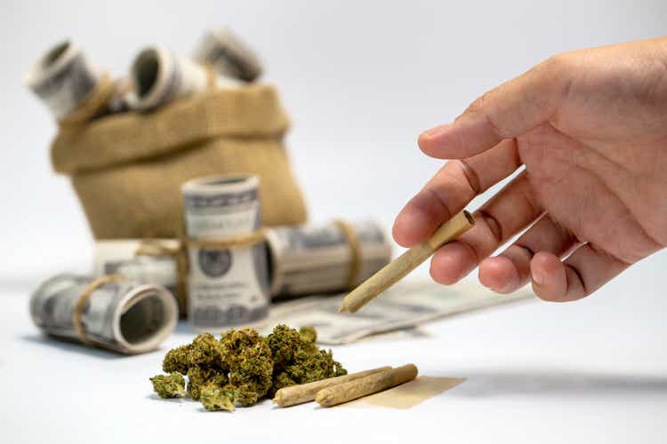 Dried marijuana and a large bag of dollar bills. Hand picked up a dried marijuana roll.