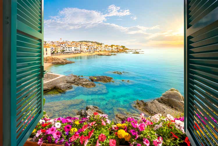 View through an open window with shutters of the sandy beach, rocky coastline and whitewashed town of Calella de Palafrugell, Spain, on the Costa Brava coast as the sun sets.