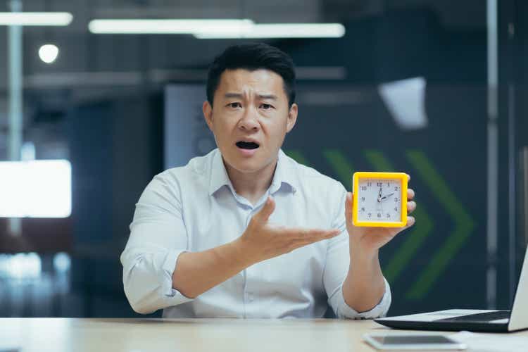 Time management. Young asian businessman holding a desk clock, pointing to the clock, confused