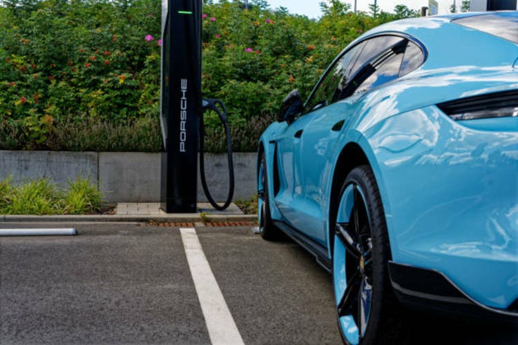side view of a blue Porsche Taycan 4S parked electric charge station with black cable. Green bush and black rim wheel.