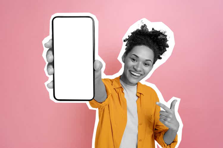 Excited happy black and white african american girl, holding smart phone in her hand with blank white mock-up screen for presentation, stands on isolated pink background, smiling. Cut out concept