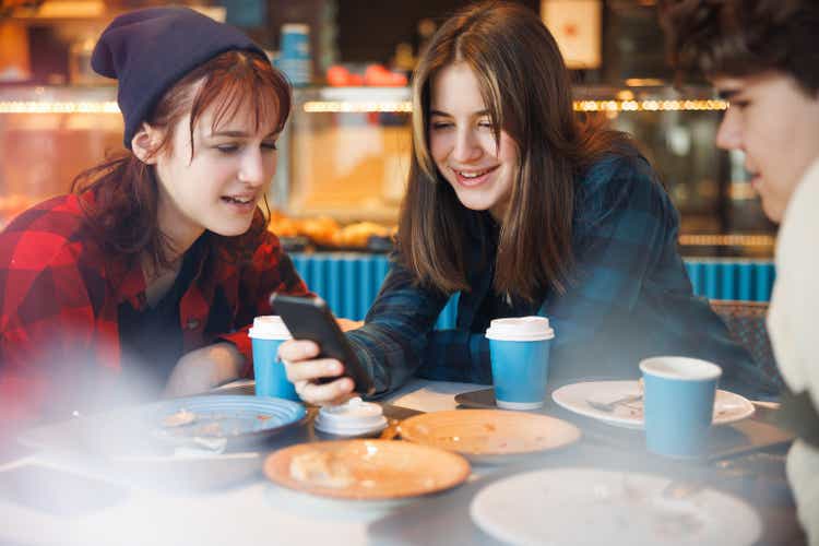 Cheerful teenage miss  having a snack with friends successful  cafeteria and utilizing astute  phone