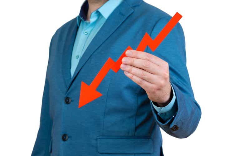 A businessman"s hand is holding a red arrow down on a white background. The concept of reducing costs and profits, falling living standards and prices. Decreased projections, depressed economies.