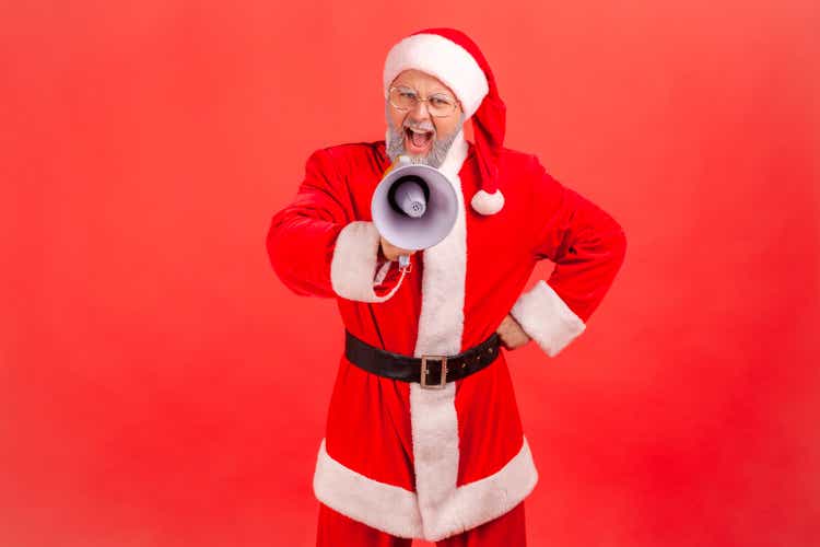 Elderly man with gray beard wearing santa claus costume screaming holding loudspeaker megaphone, having fun celebrating winter holidays.