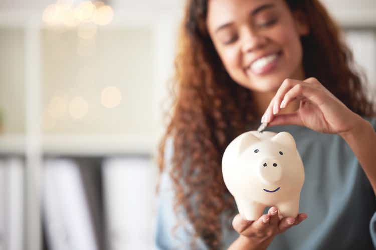 One happy young mixed race woman holding a piggybank and depositing a coin as savings. Hispanic woman budgeting her finances and investing money into her future. Saving funds for financial freedom