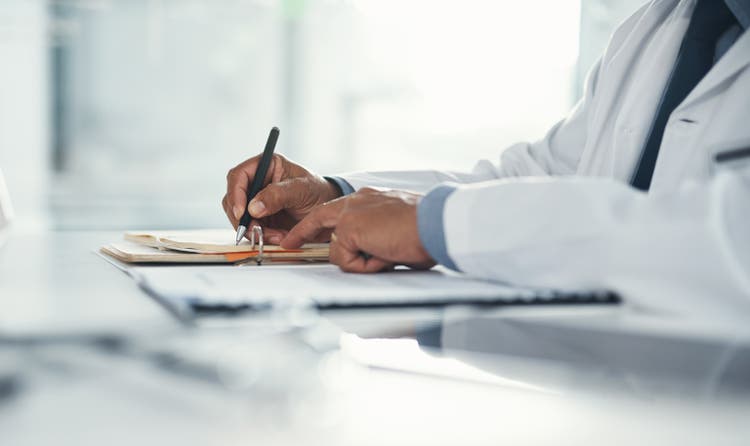 Doctor hands writing on paper or document at a desk in the hospital. Healthcare professional drafting a medical insurance letter, legal paperwork or form. A GP filing a document in a clinic office