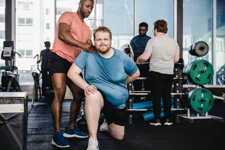 Male fitness instructor assisting overweight man in exercise at gym