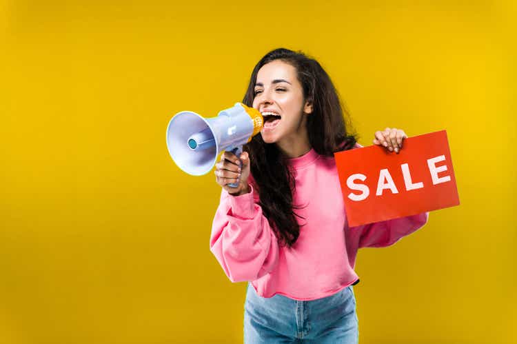 Pretty caucasian young woman emotionally shouts into a megaphone and holds a red "sale" sign in his hand standing on isolated yellow background