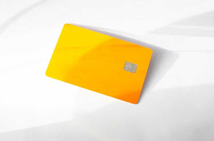 Plastic credit card with chip visible, on top of a table with soft lights and shadows. Orange color card on white surface. Concept: finance, purchases, payments, loan, spending, investments and debts.