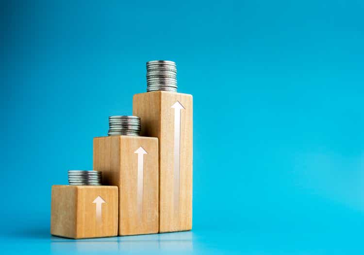 Shining rise up arrow on wooden cube blocks top with coins stacked, bar graph chart steps on blue background with space, investment, income, inflation, business growth, economic improvement concepts.