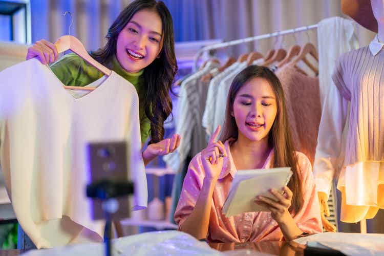 Fashion vlogger presenting dress to cellphone and showing clothes selling online live streaming ,Asian small business owner working at home studio, Live video for present clothes.