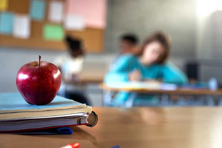 Apple and a pile of books and pens on teacher table in classroom. Focus on foreground.