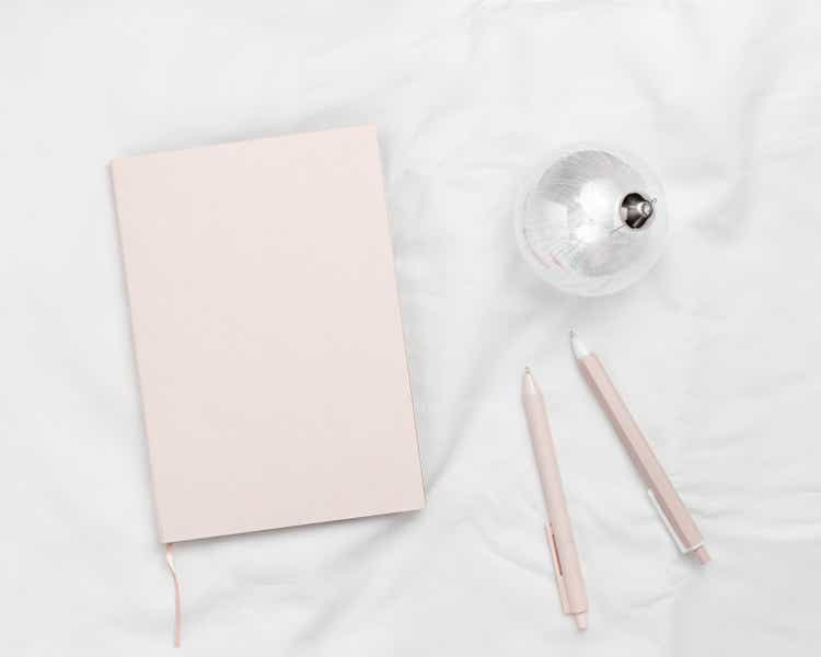 New Year planning concept, female work, pale pink coral colored diary for year 2023, pen, trend christmas glass ball with rainbow glare on white textile background. Minimalistic lifestyle.