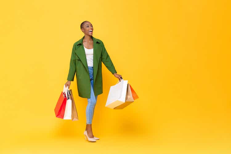 Trendy fashionable African American woman carrying colorful shopping bags walking on yellow color studio isolated background