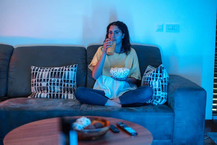 Young Latin Female Drinks Orange Juice While Watching TV. She Holds Popcorn