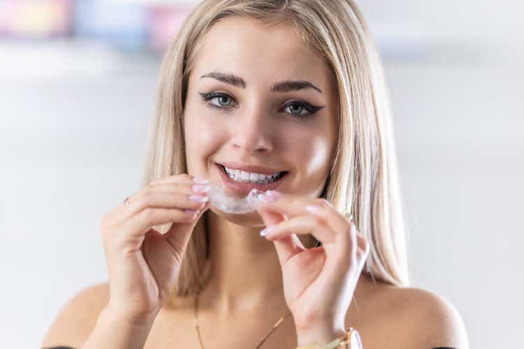 Portrait of beautiful young woman with perfect teeth holding orthodontic retainers in dental clinic.