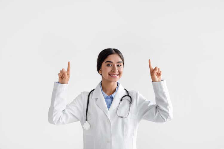 Happy millennial indian lady doctor in coat with phonendoscope raises hands and shows fingers up