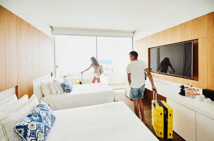 Wide shot of siblings rolling luggage into hotel room while on vacation