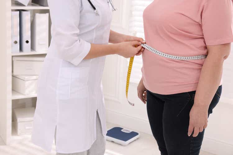 Nutritionist measuring overweight woman"s waist with tape in clinic, closeup