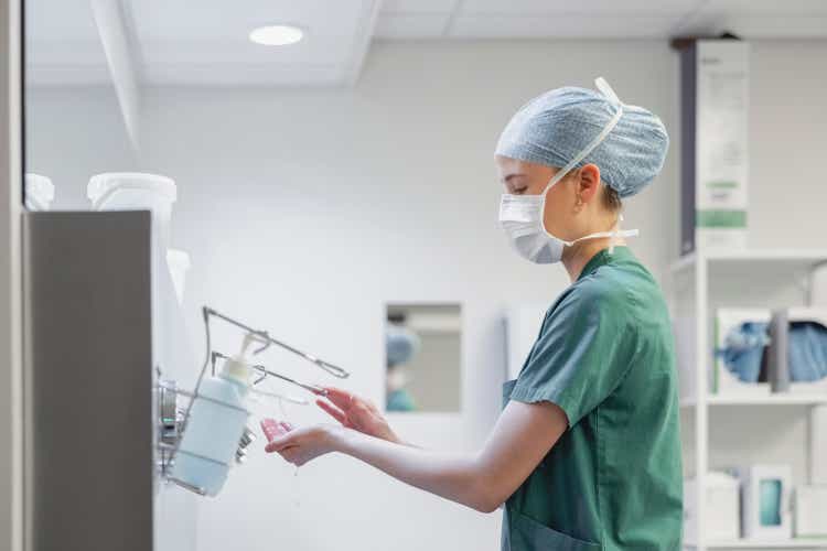 Female nurse in surgical mask and cap using hand sanitizer in operation room