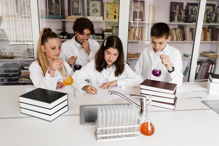 Schoolchildren studying at chemistry lesson in classrom. Pupils writing in notebook, holding flasks with liquid for experiments and have fun together. School education.