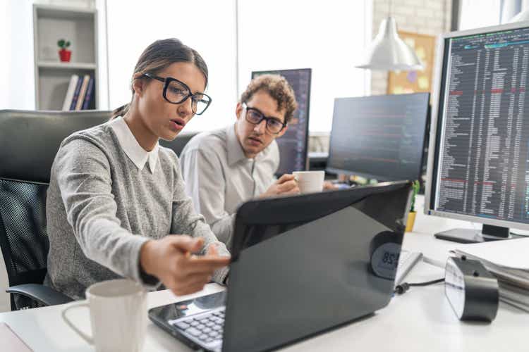Young female programmer standing on her desk, coding on a laptop, having discussion with a male colleague, giving her advice