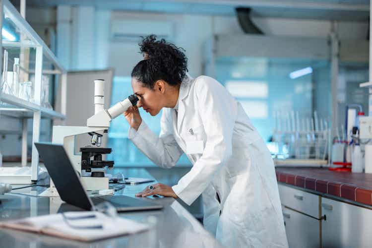 Female Scientist Looking Under Microscope And Using Laptop In A Laboratory