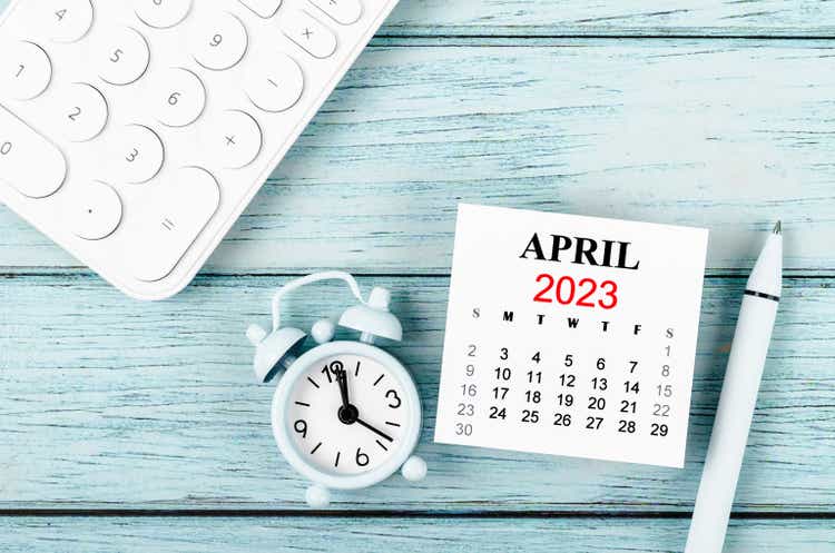 The April 2023 Monthly calendar year and alarm clock with calculator on blue wooden background.