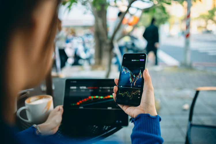 Business Woman hand using smart phone with cafe shop. Stock market charts on phone and laptop screen. Checking financial market.