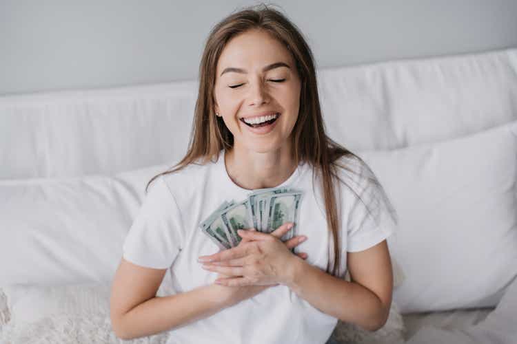 Excited blonde caucasian girl broad smiling eyes closed presses banknotes to chest, got salary, sits on bed. Happy Swedish young woman received money, celebrates. Prize, lottery, winner. Wage. Cash