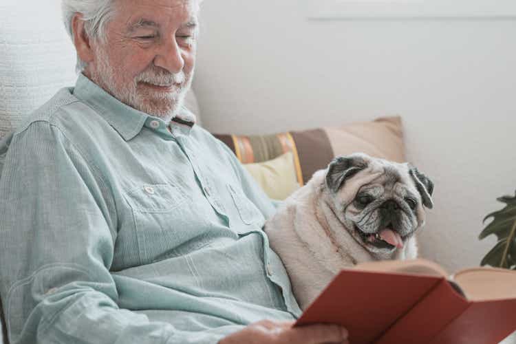 Portrait of old pug sitting on the couch at home and reading with his senior owner.Best friends and pet therapy concept