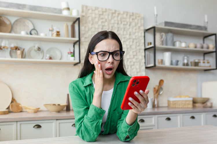 A worried and shocked young woman is standing at home in the kitchen, holding the phone in her hands