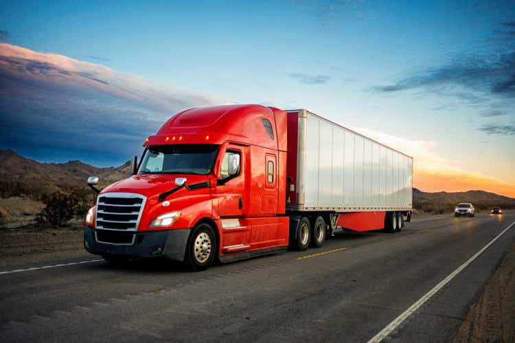 Brightly Red Colored Semi-Truck Speeding on a Two-Lane Highway with Cars in Background Under a Stunning Sunset in the American Southwest