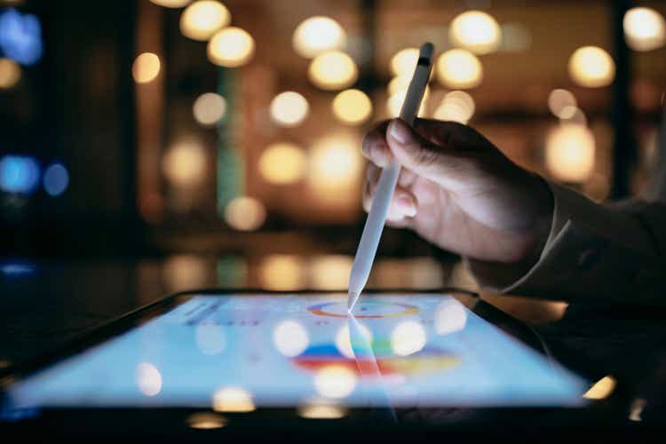 A business person is working in a cafe at night, using a tablet device and a stylus to look up chart indexes and using them for work projects.