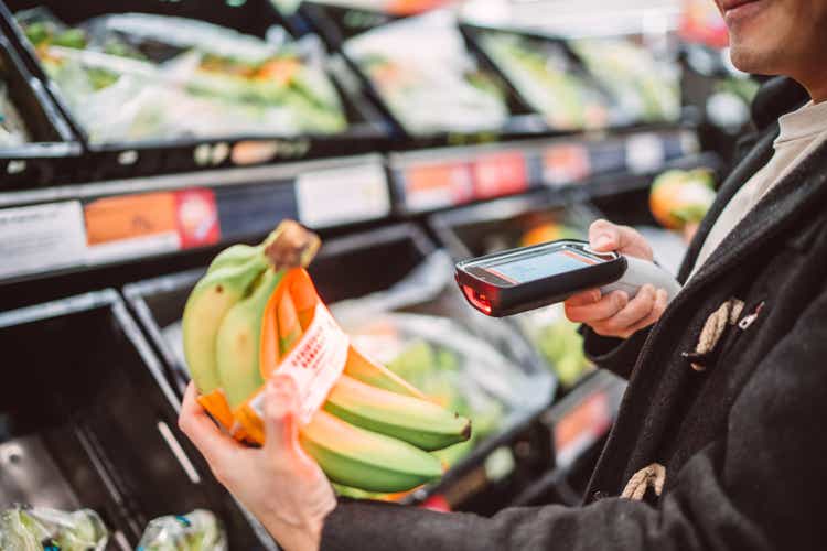 Close-up of a young man using handheld barcode reader scanning a bunch of bananas while shopping in supermarket