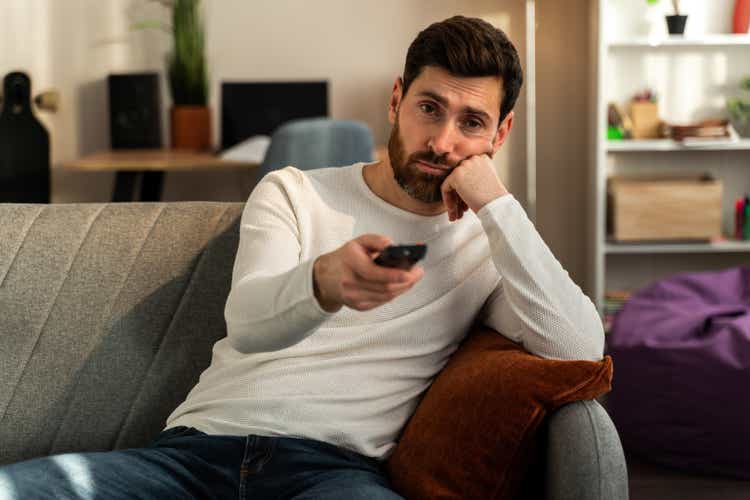 Photo of young bored man sitting on couch at home and changing TV channels with disinterest