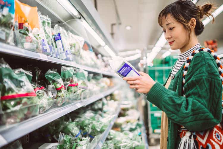 Young pretty woman checking product nutrition facts of a salad kit while shopping in supermarket