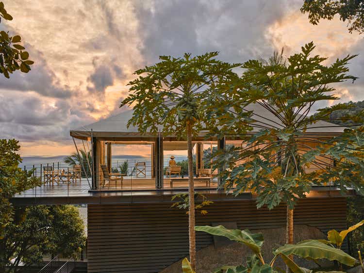 Female tourist admiring sea view while sitting on decking of tented villa, illuminated at sunrise with open sides, showing the interior. Situated on eco friendly luxury Glamping resort.