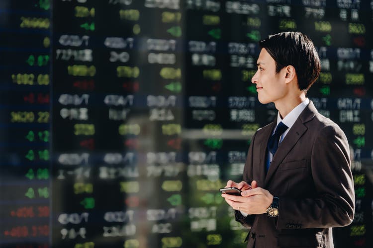 Men in their thirties in their 30s who trade with a smartphone app in front of the real -time stock price board on the electric bulletin board