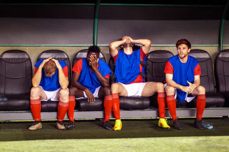 Benched professional soccer players sit with their head in their hands while watching their teammates lose