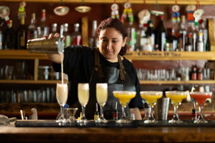 Female bartender pours another mixed drink into glass next in line to colorful cocktails at bar