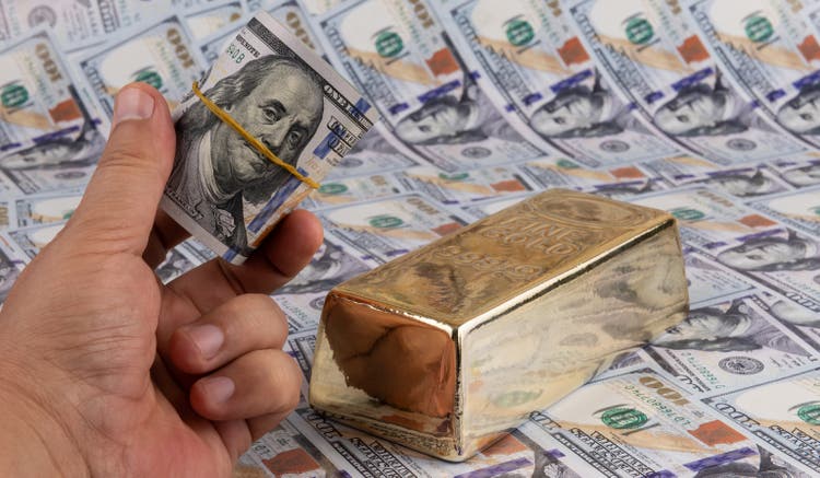 A stack of 100 amrican dollar bills in a man"s hand and a bar of gold