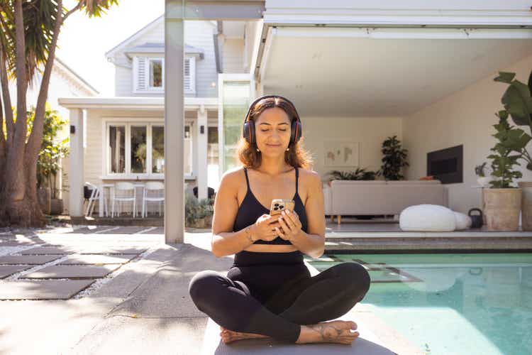 Woman with long hair, wearing headphones and exercise clothing, sitting cross-legged by the pool, using a mobile phone