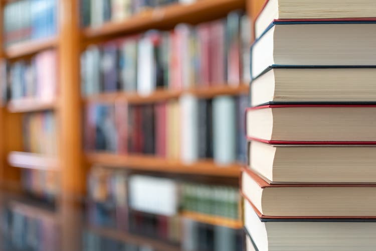 Many books stacked with blurred background of bookshelf full of books. Photo with copyspace.