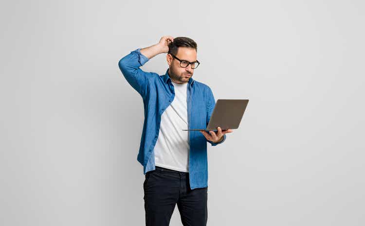 Confused disappointed businessman with hand in hair reading e-mail over laptop on gray background