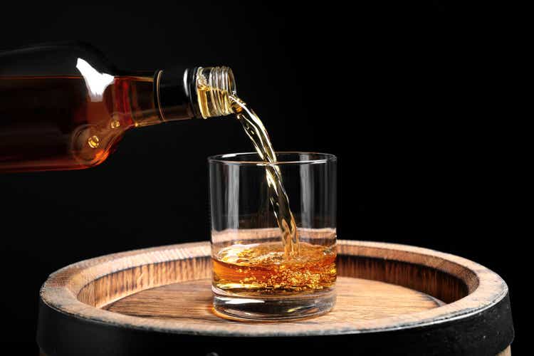 Pouring whiskey from bottle into glass on wooden barrel against black background, closeup