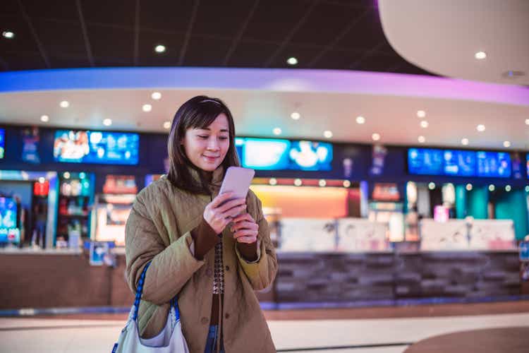 Young Asian woman using e-ticketing reservation on mobile app with her smartphone in front of ticket counter at cinema