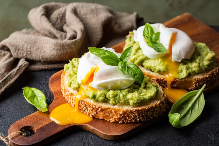 Smashed avocado sandwich with boiled poached egg with flowing yolk and basil leaves. Dark green background.