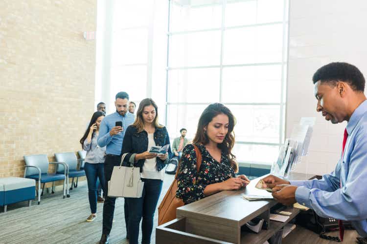 As the line grows, the bank teller counts the women.